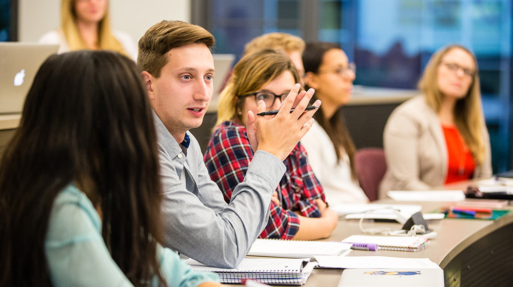 A group of people sits in a classroom, engaging in discussion. A young man in the center is speaking, gesturing with his hand. Others are listening attentively, with notebooks and laptops on the desks.